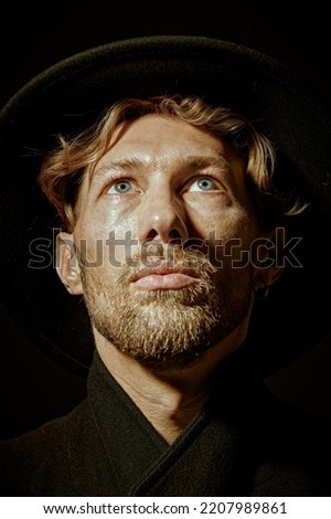 Close up portrait of a pensive mature man with beard and moustache dressed in black coat and black hat looking calmly and thoughtfully up. Black background. Male beauty. 