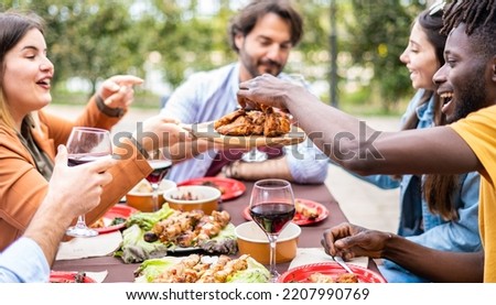 Group multi-ethnic friends having lunch at farmhouse table - Diverse young people sitting at home during bbq party -  African guy taking the grilled chicken wings by hands Royalty-Free Stock Photo #2207990769