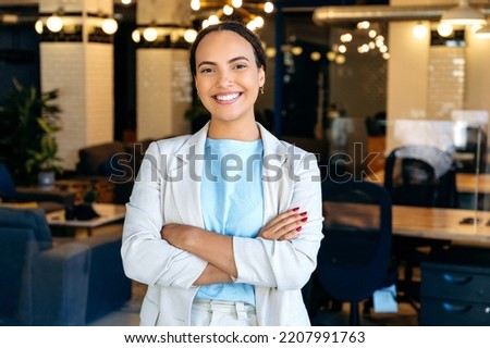 Portrait of a confident successful modern mixed race business lady, top manager of a company, standing in her modern creative office, wearing formal wear, arms crossed, looking at the camera, smiling Royalty-Free Stock Photo #2207991763