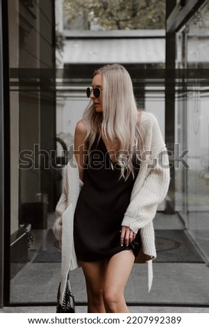 beautiful young girl dressed in black mini dress with straps, beige knitted oversized cardigan, massive boots, sunglasses, bag, accessories, wavy hair, stylish fashion outfit, lifestyle model Royalty-Free Stock Photo #2207992389