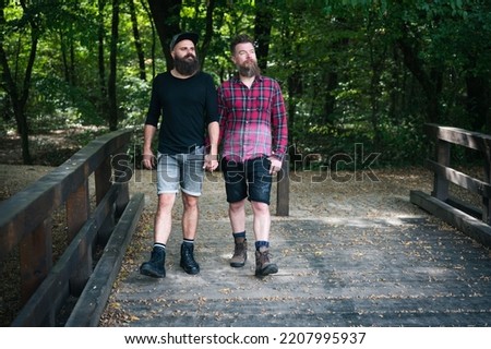 Long bear hipster gay couple walking in park toward camera, crossing wooden bridge and holding hands. Good looking homosexuals outdoor in autumn.  Royalty-Free Stock Photo #2207995937
