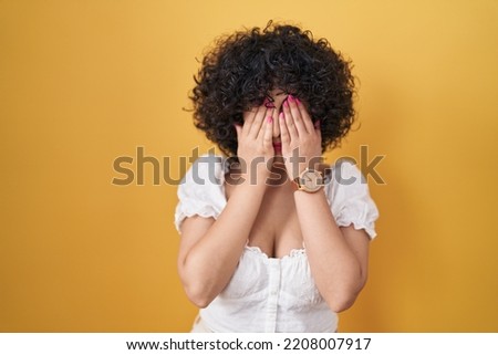 Young brunette woman with curly hair standing over yellow background rubbing eyes for fatigue and headache, sleepy and tired expression. vision problem 