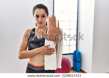Young brunette woman wearing sportswear and towel at the gym doing stop sing with palm of the hand. warning expression with negative and serious gesture on the face. 