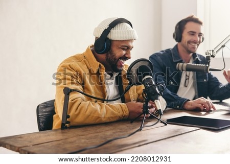 Two college podcasters laughing and having a good time in a studio. Two happy young men co-hosting a live audio broadcast. Two male content creators recording an internet podcast.