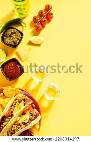 Mexican tequila shots with lime and hot red chili on the yellow table with traditional food corn tacos on the background. Top view. Copy space.