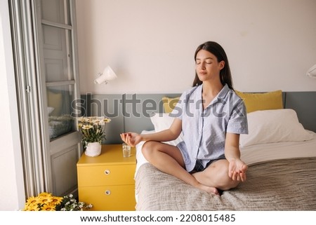 Calm down young caucasian girl with closing eyes meditating sitting on bed at home. Brunette hair woman dressed in cozy pajamas. Concept meditation, morning. Royalty-Free Stock Photo #2208015485