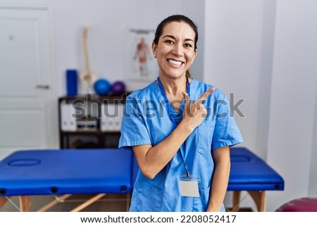 Young hispanic woman wearing physiotherapist uniform standing at clinic cheerful with a smile of face pointing with hand and finger up to the side with happy and natural expression on face 