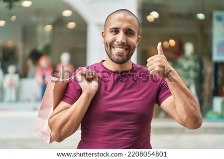 Hispanic man with beard holding shopping bags outdoors smiling happy and positive, thumb up doing excellent and approval sign 
