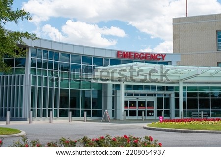 A medical hospital's emergency department sign is made of red glass. The sign is attached to a clear glass building overhang. There are trees and blue sky in the background. The sign is not lit up. Royalty-Free Stock Photo #2208054937