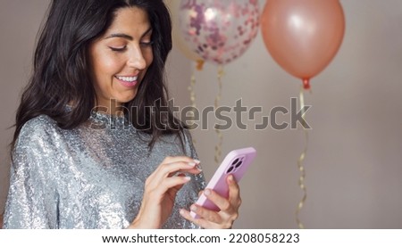 Beautiful smiling  woman with shiny dress using her mobile phone  on balloons background .Party time concept 