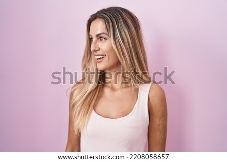 Young blonde woman standing over pink background looking away to side with smile on face, natural expression. laughing confident. 