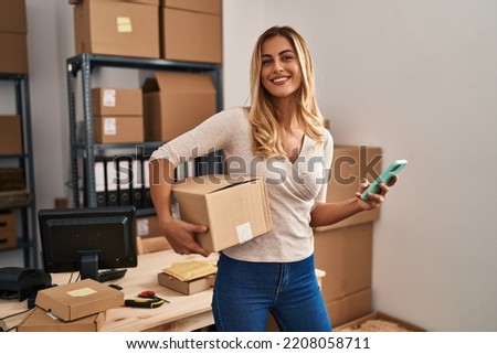 Young blonde woman ecommerce business worker using smartphone holding package at office Royalty-Free Stock Photo #2208058711
