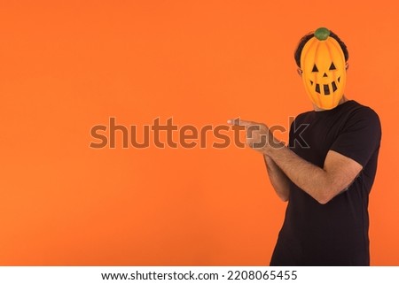 Person with pumpkin mask celebrating Halloween, pointing to the side with fingers, on orange background. Concept of celebration, All Souls' Day and All Saints' Day.