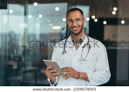 Portrait of happy and successful african american doctor man working inside office clinic holding tablet computer looking at camera and smiling wearing white coat with stethoscope Royalty-Free Stock Photo #2208078001