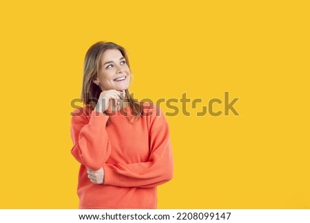 Happy young woman thinking with hand on chin and positive expression. Creative cheerful confident modern teenage girl in orange jumper dreaming of something good on vivid yellow copyspace background