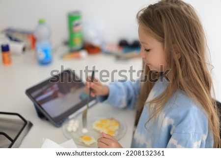 Positive caucasian little girl having breakfast and looking at tablet at home