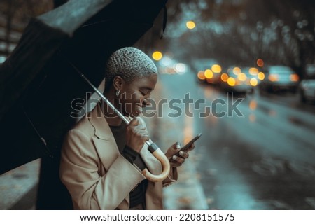 A profile view of a young elegant cheerful black lady with short painted white hair, using her umbrella on the rainy street while trying to call a taxi via an app on her smartphone to escape the rain Royalty-Free Stock Photo #2208151575