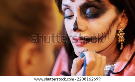 Halloween party, make-up artist draws a terrible makeup on the face of a brunette woman for a Halloween party. in the background, the scenery in the style of Halloween is seen. High quality photo