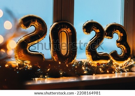 2023 golden foil balloons on blue window sill. Celebrating holidays at home, festive decor concept. Happy New Year 2023. close-up numbers of year 2023 on dark background. Bokeh warm garland light.