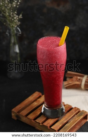 red fruit juice in a glass with a straw on a black background and wooden textures, flowers behind