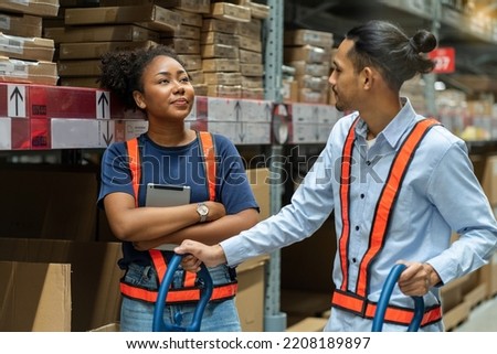 Asian male employee walks a shopping cart greets an African-American female employee sorting goods on the shelf, chatting while working in a warehouse.