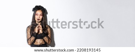 Thoughtful and cunning asian woman in witch costume prepared something for halloween, looking upper left corner and thinking. Girl wearing black gothic dress and wreath for party
