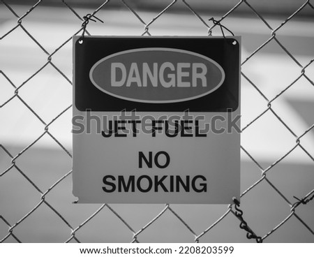 Black and white photograph of a Danger, Jet Fuel, No smoking sign on a fence. 
