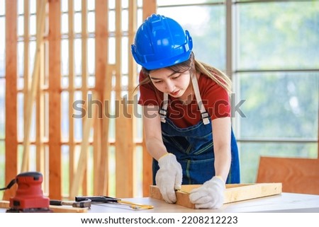 Asian professional female engineer architect foreman labor worker wears safety hard helmet jeans apron and gloves standing using squard angle degree ruler and pencil measuring wood plank on workbench. Royalty-Free Stock Photo #2208212223