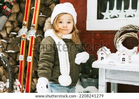 cute little girl in knitted white hat stand at porch of country house decorared for celebration New Year and Christmas holidays for children outdoor