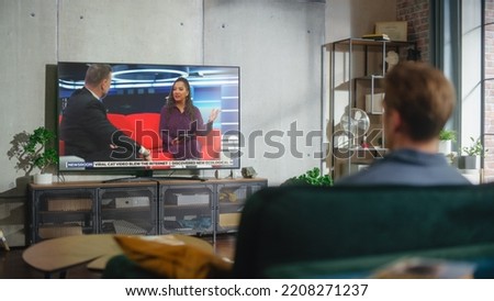 Young Handsome Man Sitting in Relaxed Pose on a Sofa and Watching TV with Live Talk Show During the Day Time on Weekend. Serious Female African American Host Interviews Guest in Studio. Royalty-Free Stock Photo #2208271237