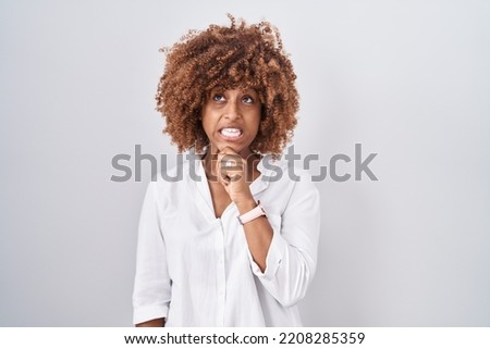 Young hispanic woman with curly hair standing over white background thinking worried about a question, concerned and nervous with hand on chin 