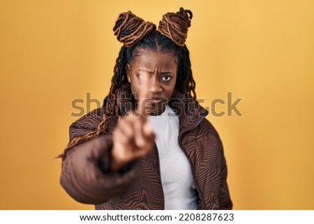 African woman with braided hair standing over yellow background pointing with finger up and angry expression, showing no gesture 