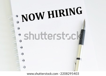 Now Hiring Text on a Sheet of Notepad on a Bright Table Next to A Pen, Business Concept