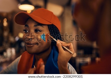 African American sports fan painting heart shape in team's colors on her face while getting ready for the match during soccer world cup.  Royalty-Free Stock Photo #2208299301