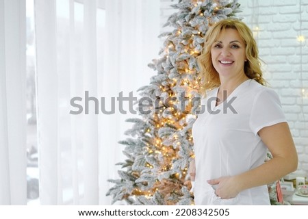 A beautiful female doctor in a white medical suit stands against the backdrop of a Christmas tree, smiling and looking at the camera. Happy family holidays medical staff. Horizontal photo