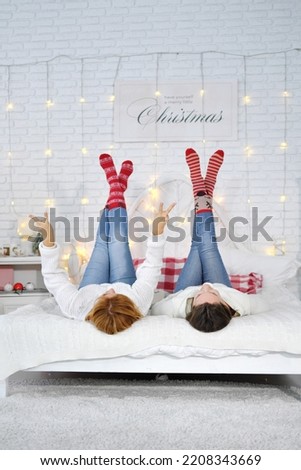 friendship and christmas home party concept - two young female friends lying down on the bed with legs up in Christmas striped socks. Vertical photo