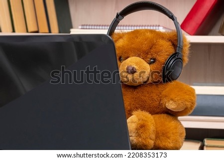 Toy teddy bear wearing headphones sitting at his laptop against a background of shelves of books and documents, close-up, selective focus. Concept: remote work, communication via the Internet.
