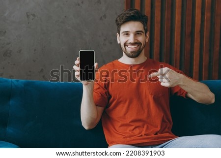 Young man wears red t-shirt hold use point finger on mobile cell phone blank screen workspace sit on blue sofa stay at home hotel flat rest relax spend free spare time in living room indoors grey wall