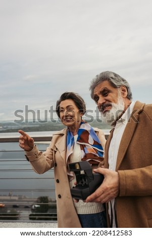 happy senior man holding vintage camera near wife smiling and pointing with finger on bridge
