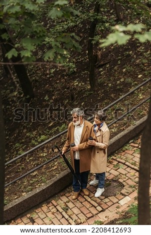 high angle view of pleased and retired couple in beige coats holding paper cups while walking in park