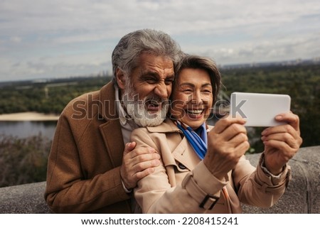 happy senior woman in trench coat and taking selfie with bearded husband