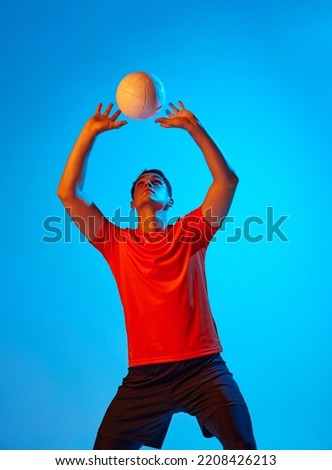 One male volleyball player in orange-black uniform training with ball isolated on blue studio background in neon light. Sport, gym, team sport, challenges. Athlete playing volleyball