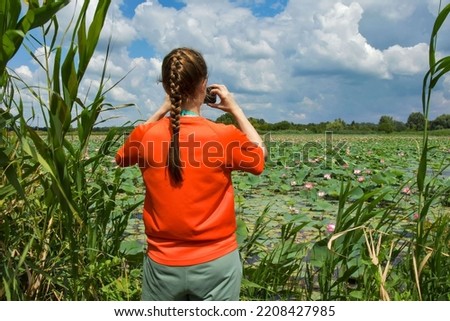 A girl in bright orange clothes shoots a beautiful lake with lotuses on a smartphone. Place for text. A lone tourist photographs a pond overgrown with water lilies.