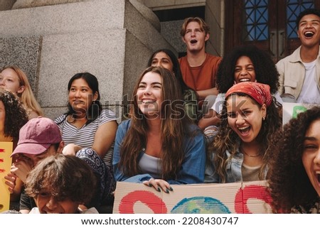 Group of youth activists smiling happily while sitting outside a building with posters and banners. Multicultural young people protesting against global warming and climate change. Royalty-Free Stock Photo #2208430747