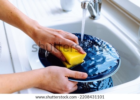 Women's hands wash dirty plate with sponge for dishes under stream of water from tap. concept of cleaning dirty dishes after eating, household chores, kitchen sink, water consumption Royalty-Free Stock Photo #2208431101