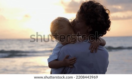 Close-up against the background of the setting sun in the sunset, on the ocean, is a woman in her arms with a child. She kisses the baby and hugs her tightly.