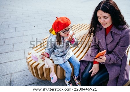 Brunette caucasian positive woman and daughter using smartphone sitting on the bench at city street. Mother calms the girl showing cartoon on the phone. Happy family moments in the city.