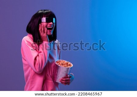 Overjoyed excited pretty brunet lady in pink hoodie specular sunglasses with popcorn open mouth touch cheek posing isolated in blue violet color light background. Neon party Cinema concept. Copy space