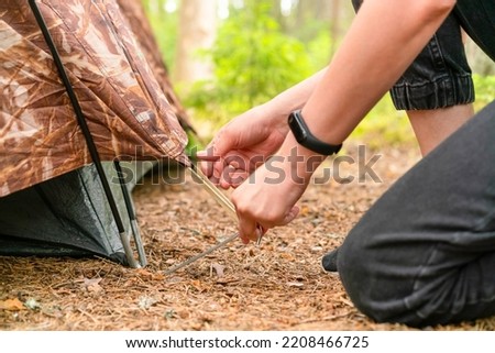 Close-up of a woman's hand. Pulls and sticks a metal tent peg into the ground. Setting up a tent in a coniferous forest. Blurred foreground. Royalty-Free Stock Photo #2208466725