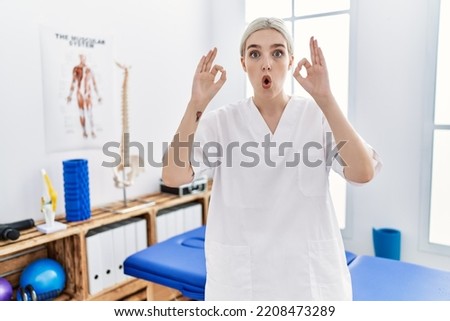 Young caucasian woman working at pain recovery clinic looking surprised and shocked doing ok approval symbol with fingers. crazy expression 